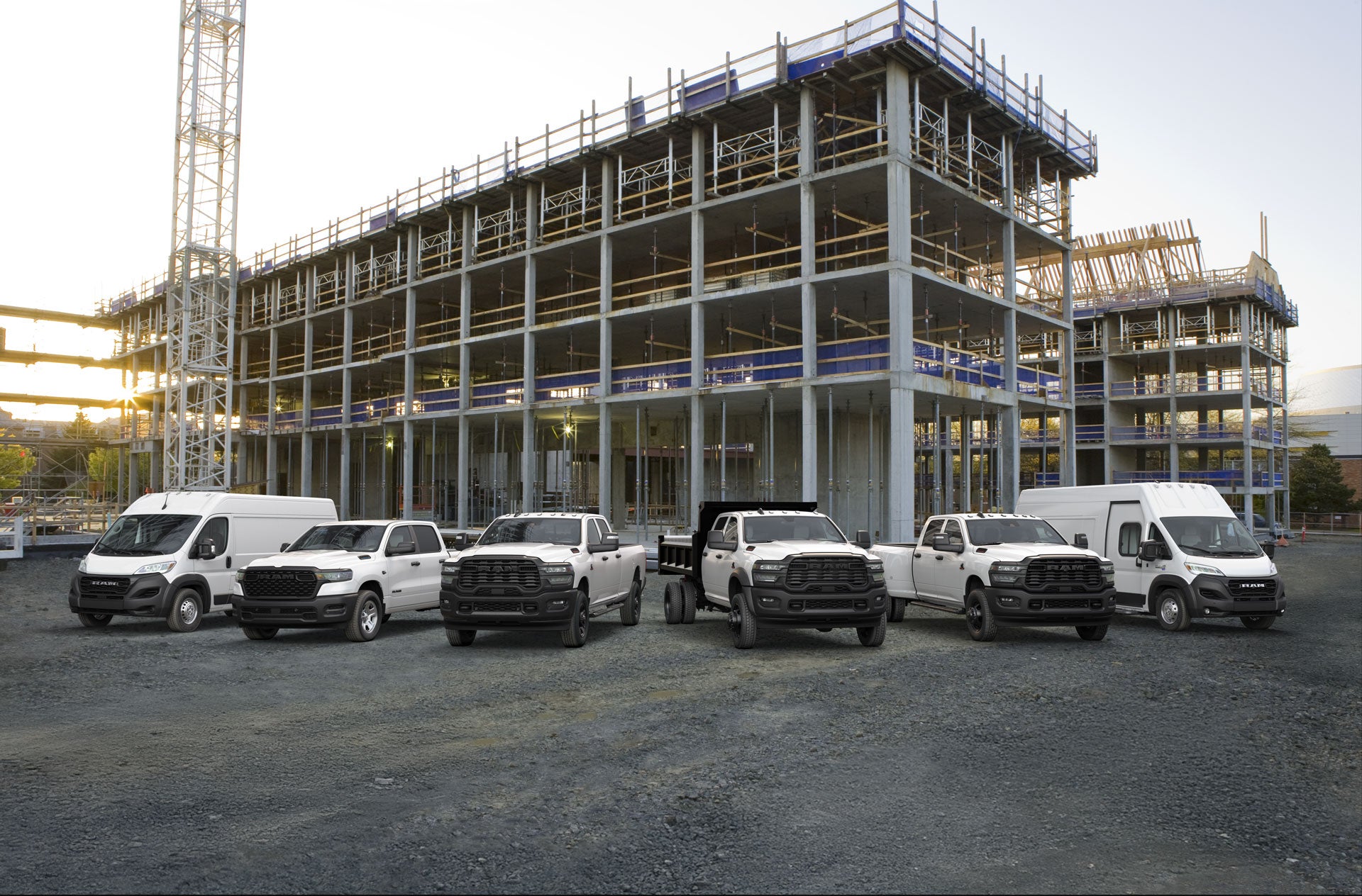 A fleet of white Ram trucks and vans parked on a gravel lot in front of a concrete building under construction.